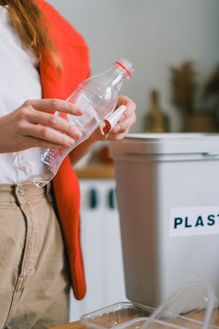 Close-up of a person wearing a white shirt and beige trousers, holding an empty clear plastic water bottle with a red cap, preparing to dispose of it into a recycling bin labeled 'PLASTIC' in capital letters. The scene is set indoors, likely in a kitchen or laundry room, with a countertop and other household items visible in the background, including a blurred appliance. The person’s hand is grasping the bottle, and the background is slightly out of focus to emphasize the recycling activity. This image relates to proper waste disposal as part of home relocation or packing processes handled by Man with Van Wallington, aiding in efficient house removals and moving logistics.
