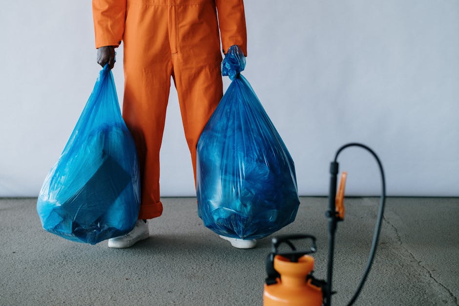 Close-up of a person dressed in orange work overalls standing on a concrete floor, holding two large blue plastic bags filled with waste or materials for house clearance, with their hands at their sides. The person's face is not visible in the image. A utility sprayer or pressure washer with an orange tank and black hose is positioned on the floor in the foreground. The background features a plain white wall, and the setting appears to be indoors, likely in a property prepared for moving or debris removal, with the scene emphasizing the process of waste collection and packing prior to a house removal in Wallington, as managed by Man with Van Wallington and their removals services.