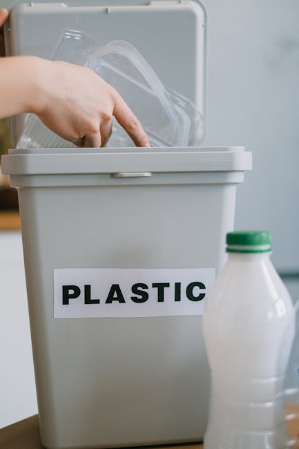 Close-up of a person wearing a white shirt and beige trousers, holding an empty clear plastic water bottle with a red cap, preparing to dispose of it into a recycling bin labeled 'PLASTIC' in capital letters. The scene is set indoors, likely in a kitchen or laundry room, with a countertop and other household items visible in the background, including a blurred appliance. The person’s hand is grasping the bottle, and the background is slightly out of focus to emphasize the recycling activity. This image relates to proper waste disposal as part of home relocation or packing processes handled by Man with Van Wallington, aiding in efficient house removals and moving logistics.
