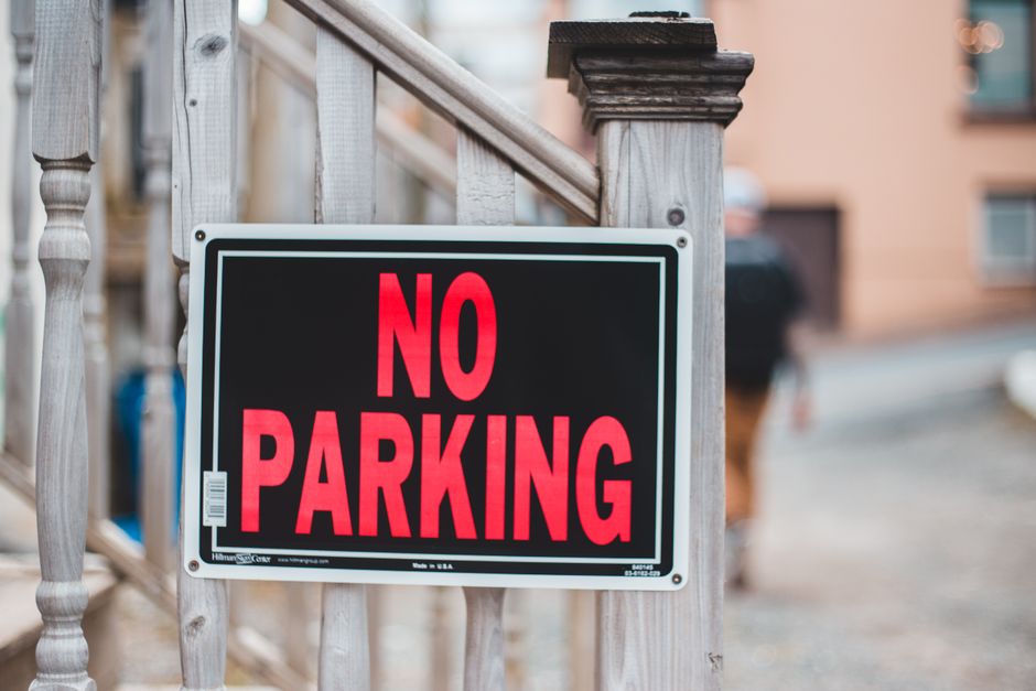 A close-up image of a rectangular 'NO PARKING' sign attached to a wooden railing outside a residential property. The sign features bold red capital letters on a black background with a thin white border. Behind the sign, part of a wooden staircase with vertical balusters and a newel post is visible, indicating access to the house entrance. The background shows a blurred view of a street with parked cars and residential buildings, suggesting an urban or suburban neighbourhood. The lighting appears natural, typical of daytime. This scene relates to house relocations, as clear parking restrictions like this can impact the logistics of furniture transport and moving processes, highlighting the importance of planning for parking during home removal services and the need for professional assistance from companies like Man with Van Wallington.