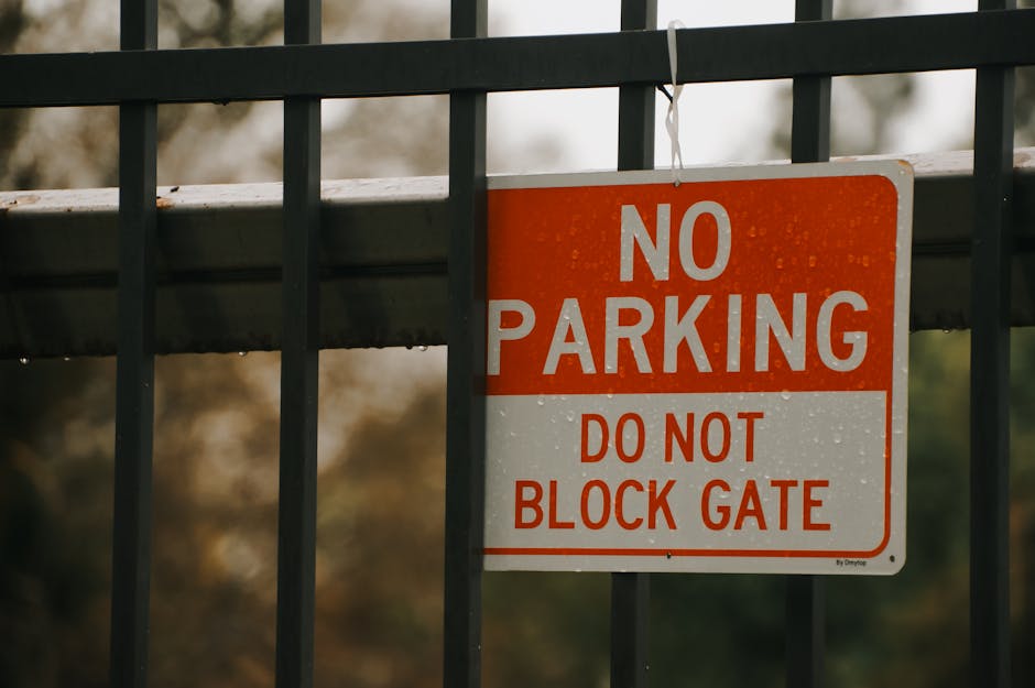 A rectangular metal sign attached to a black iron fence with vertical bars, featuring a red and white colour scheme. The sign displays the message 'NO PARKING' in bold white letters on a red background at the top, followed by 'DO NOT BLOCK GATE' in red text on a white background below. The fence is positioned on a pavement near a gate, with the sign hanging from the fence. Behind the fence, blurred trees and foliage are visible, indicating an outdoor setting. The scene suggests a location where parking restrictions are enforced, relevant to house removals and moving logistics conducted by Man with Van Wallington during home relocation or furniture transport processes.
