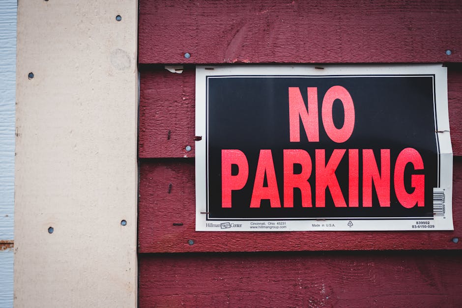 A close-up image of a rectangular 'NO PARKING' sign attached to a wooden railing outside a residential property. The sign features bold red capital letters on a black background with a thin white border. Behind the sign, part of a wooden staircase with vertical balusters and a newel post is visible, indicating access to the house entrance. The background shows a blurred view of a street with parked cars and residential buildings, suggesting an urban or suburban neighbourhood. The lighting appears natural, typical of daytime. This scene relates to house relocations, as clear parking restrictions like this can impact the logistics of furniture transport and moving processes, highlighting the importance of planning for parking during home removal services and the need for professional assistance from companies like Man with Van Wallington.