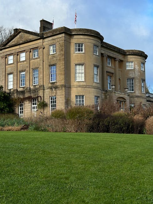 A large, historic stone building with multiple windows, including some with small panes and others with larger glass sections, set against a clear blue sky with minimal clouds. The building features rounded corner sections and a flat roof with a small flagpole and the Union Jack flying. Surrounding the structure is well-maintained greenery, including shrubs and grass, with a neatly trimmed lawn in the foreground. The scene suggests a stately property suitable for house moves or relocation services. The building’s exterior appears weathered, indicating age and traditional architecture, and is likely an estate or institutional building. The overall environment is calm and professional, fitting for a house removal or furniture transport context, as would be managed by companies like Man with Van Wallington, especially when preparing for packing and moving activities. The image emphasizes the scale of the property and its suitability for home relocation services in Wallington.