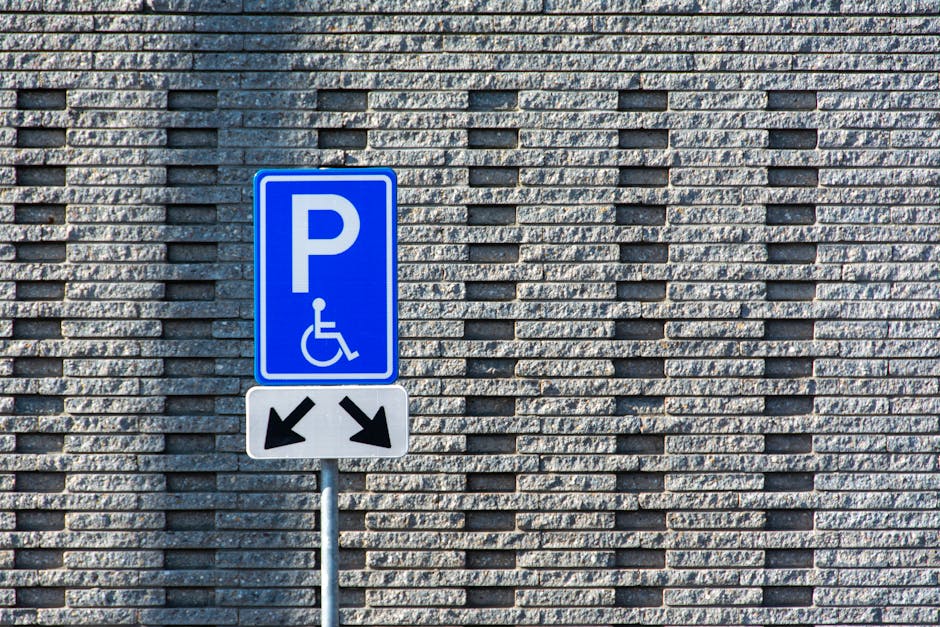A blue parking sign with a white 'P' symbol and a wheelchair icon indicating designated accessible parking is mounted on a metal pole in front of a textured grey brick wall. The sign also features two black downward-pointing arrows, one on each side. The scene is outdoors with natural lighting, and the sign is situated at a typical height for visibility. This parking signage is relevant to house removals and moving services provided by Man with Van Wallington, as it highlights accessible parking options in areas where home relocation or furniture transport may require parking considerations leading up to entry points of residential properties near Wallington Station.