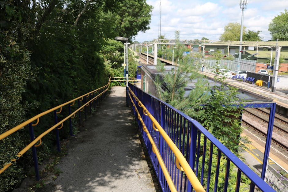 A pathway beside a railway station with a blue metal railing on the right side and a yellow handrail on the left, leading towards a small metal gate. The pathway is paved with asphalt and partially shaded by overhanging trees on the left. In the distance, railway tracks run parallel to the platform, which has benches, signage, and parked cars visible across the tracks. The scene is outdoors under a partly cloudy sky, indicating daytime. This setting reflects a typical environment involved in home relocation or furniture transport, as part of a house removal process, supported by Man with Van Wallington’s services for efficient moving near Wallington Station.