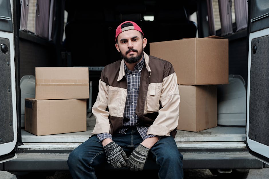 A young man with a neatly trimmed beard and wearing a red baseball cap, sitting on the edge of an open van hatchback during a home relocation process. He is dressed in a checkered shirt layered with a