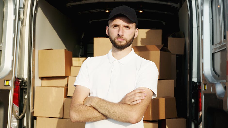 A young man with a neatly trimmed beard and wearing a red baseball cap, sitting on the edge of an open van hatchback during a home relocation process. He is dressed in a checkered shirt layered with a