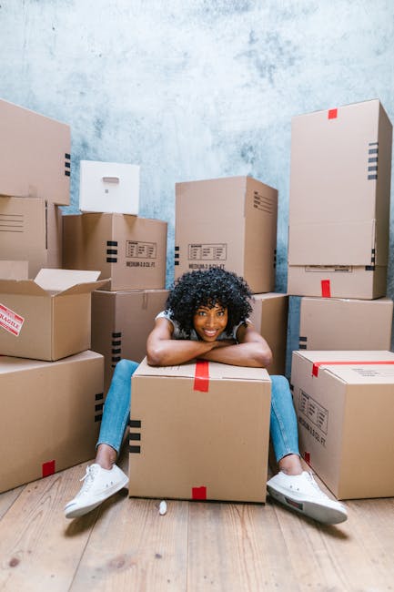 A young woman with curly black hair, wearing a white t-shirt, blue jeans, and white sneakers, sitting on the wooden floor amidst numerous cardboard boxes in a room with a plain, light-colored wall. Some boxes are arranged upright while others are lying on their sides, with visible red packing tape and labels. She is smiling and resting her arms on a large box in front of her, with her legs spread out around it. The boxes vary in size and are likely used for packing household items as part of a home relocation process. The room appears well-lit, suggesting daytime, and the scene captures the packing stage of a house move, with a focus on furniture and box transportation logistics often handled by services like those offered by Man with Van Wallington.