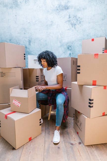 A young woman with curly black hair, wearing a white t-shirt, grey jeans, and white sneakers, is sitting on a wooden floor surrounded by large cardboard moving boxes, some sealed with red packing tape and others open revealing packing materials. She is smiling and holding a sealed box in her lap, with her left arm resting on top of it. Behind her, multiple stacked boxes are positioned close to a blue-painted wall with a slightly textured surface, indicating an indoor setting possibly within a property prepared for a home relocation. The boxes vary in size, and some are labeled with printed text and barcodes. The scene depicts the packing and loading process associated with house removals, reflecting the activities involved in furniture transport and moving logistics. This image captures a moment during packing or unpacking, relevant to services offered by Man with Van Wallington as part of their removals and relocation solutions.