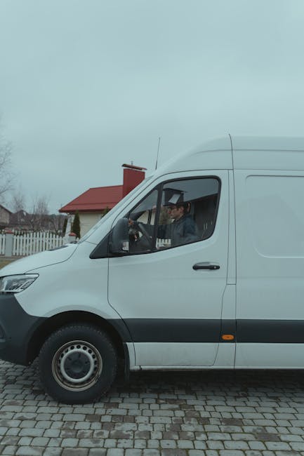 A white van with a black front bumper and side mirrors parked on a cobblestone driveway in a residential area with houses and well-maintained gardens visible in the background. The van's driver is par