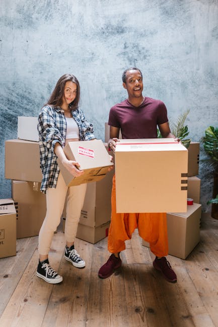 Two individuals are inside a room with a textured, light grey wall and wooden flooring, engaged in a home relocation process. The woman on the left, dressed casually in beige trousers, a white t-shirt