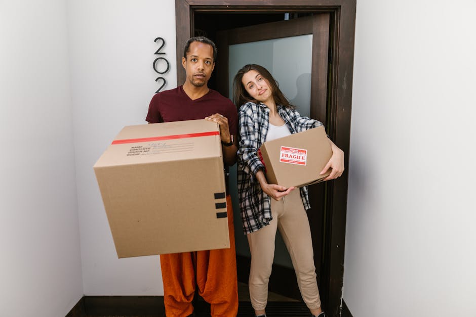 Two individuals are inside a room with a textured, light grey wall and wooden flooring, engaged in a home relocation process. The woman on the left, dressed casually in beige trousers, a white t-shirt