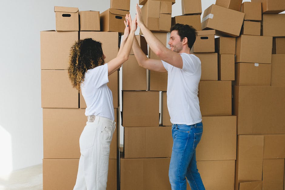 A man and a woman, both dressed in casual white t-shirts and jeans, are inside a residential property during a house removal process, standing in front of a large stack of cardboard boxes. The boxes a