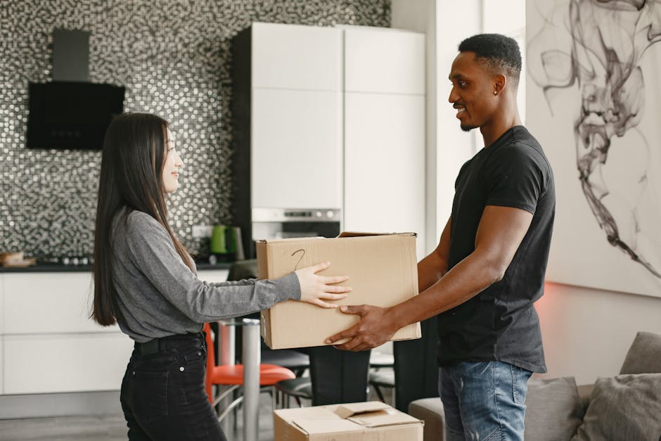 A man and a woman, both dressed in casual white t-shirts and jeans, are inside a residential property during a house removal process, standing in front of a large stack of cardboard boxes. The boxes a