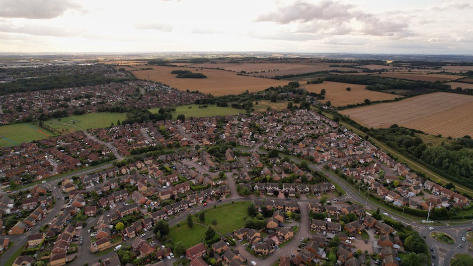 Aerial view of a suburban residential neighbourhood with numerous detached and semi-detached houses, many with driveways and gardens, arranged along curved streets. The area features a central green space with trees and grass, surrounded by rows of houses with red and brown tiled roofs. In the background, there are open fields and farmland extending to the horizon, under a cloudy sky. The image showcases the community layout typical of home relocation and furniture transport processes, with visible rooftops and streets indicating the area serviced by house removals companies like Man with Van Wallington, supporting local moving and packing services in Sutton and Wallington.