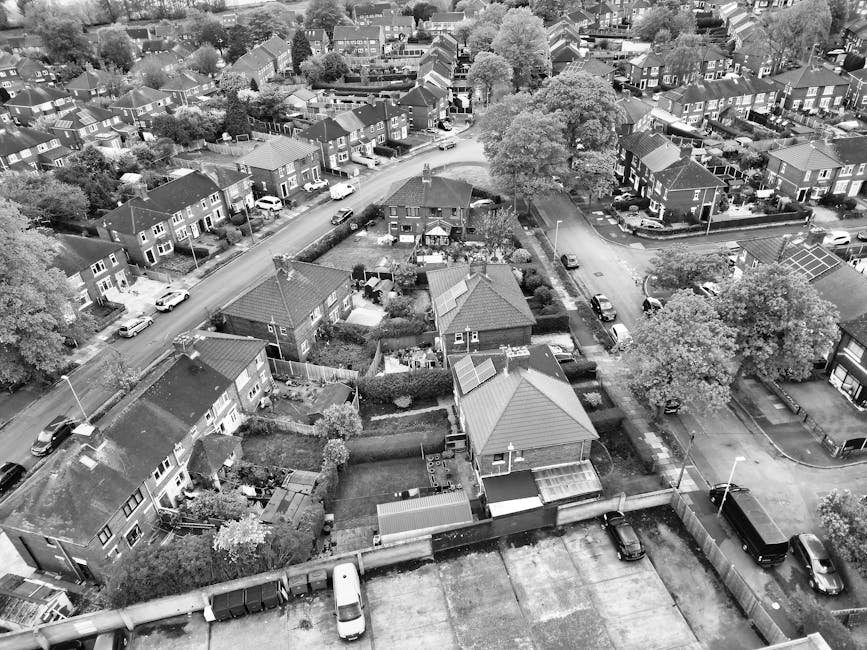 Aerial view of a suburban residential neighbourhood with numerous detached and semi-detached houses, many with driveways and gardens, arranged along curved streets. The area features a central green space with trees and grass, surrounded by rows of houses with red and brown tiled roofs. In the background, there are open fields and farmland extending to the horizon, under a cloudy sky. The image showcases the community layout typical of home relocation and furniture transport processes, with visible rooftops and streets indicating the area serviced by house removals companies like Man with Van Wallington, supporting local moving and packing services in Sutton and Wallington.