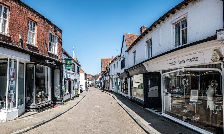 A quiet street scene on Wallington High Street during daytime, with small retail shops occupying both sides of the narrow, cobblestone road. The storefronts include a craft shop displaying handmade items in large window displays, with signage that reads 'I made this crafts,' and another shop with large glass windows showcasing various goods. The street is largely empty, with no visible pedestrians or vehicles, and the bright blue sky overhead suggests good weather. The image captures the historic, quaint character of the high street, with brick and white-painted building facades and traditional roofing visible. This setting is suitable for illustrating aspects of home relocation or furniture transport, such as the need for careful logistics and planning when moving items through tight, urban streets. Man with Van Wallington, a professional removals service, could provide expertise in managing such urban moving challenges, including loading procedures and transport logistics for residential moves along narrow streets like these.