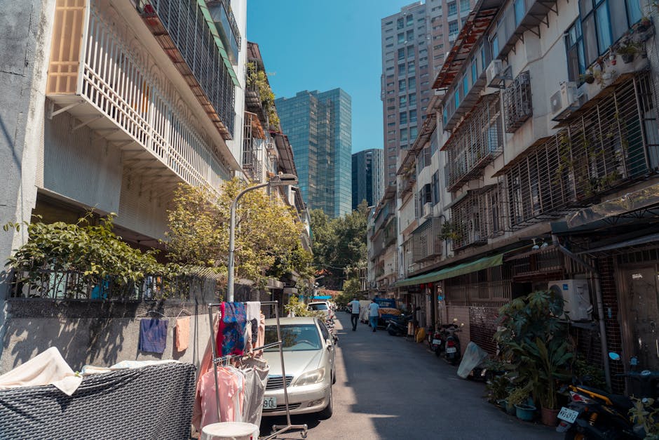 A narrow urban street at Wallington High Street during daylight, showing a mix of small retail shops and businesses housed in brick and painted buildings. Several people are gathered on the left sidewalk, some waiting or conversing near store entrances, while others walk along the pavement. Two white vans are parked on the right side—one appears to be a moving company vehicle with a partially visible logo—potentially involved in loading or unloading furniture and boxes for a home relocation. The pavement is composed of brick and concrete, with a central roadway of paved cobblestones, facilitating vehicle access and pedestrian movement. Street lamps with black metal fixtures line both sides of the street, and a red customer service point sign is visible on the right sidewalk. The scene suggests active logistics, with items being prepared or transported, and the environment includes typical small-town shopping street elements, supportively reflecting the context of furniture transport, packing, and moving activities associated with house removals, as exemplified in the Wallington high street moving guide.