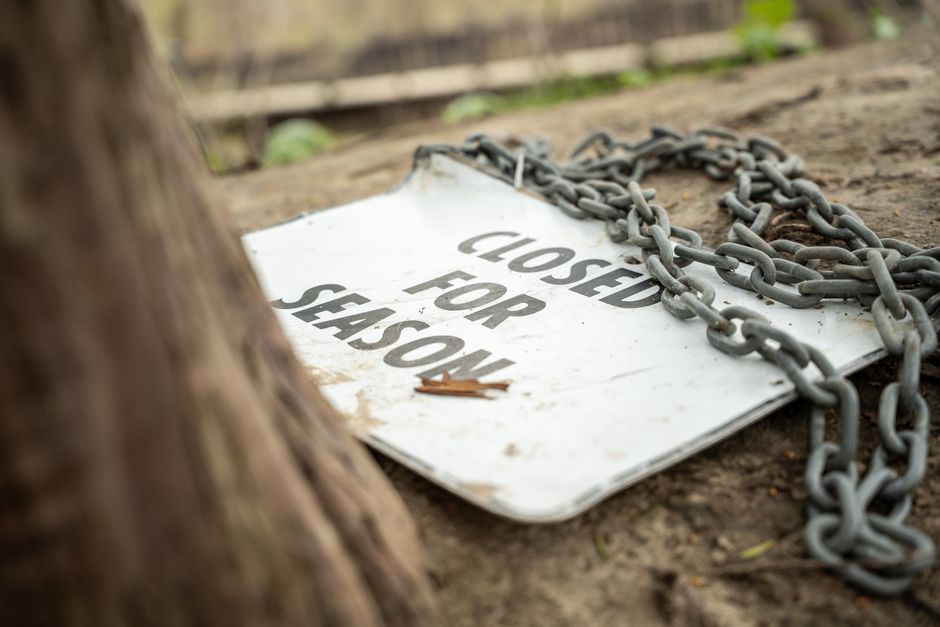 A rectangular metal sign with a white background and black text reading 'CLOSED FOR SHEDOWN' lies on the ground outdoors, partially covered by a heavy metal chain coiled on top of it. The sign appears weathered with some dirt and a small leaf resting on its surface. Adjacent to the sign, there is dirt and mud, and in the foreground, a blurred wooden fence post is visible, indicating the scene is in an outdoor setting near a property boundary. The environment suggests a temporary closure or restriction, possibly related to house removals or site access, with natural lighting illuminating the scene. This image, associated with the website manwithvanwallington.co.uk, visually captures aspects of moving logistics, such as site clearance or restricted access during a home relocation process.